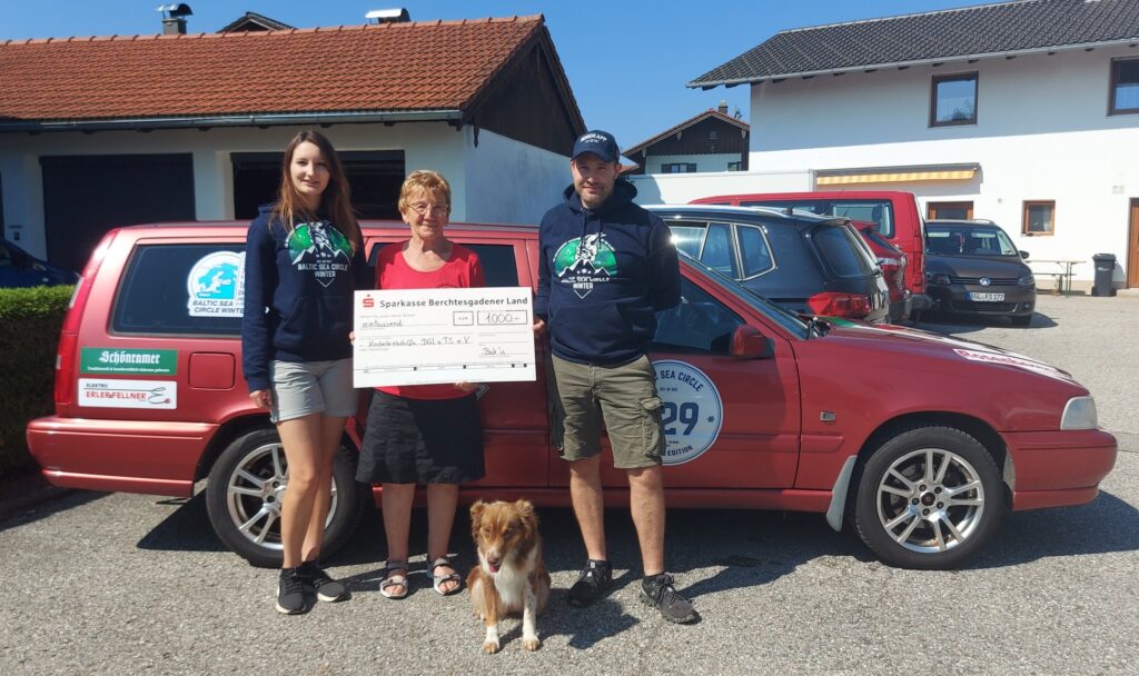 Barbara, Andi und Hund übergeben ihre Spende an Rosmarie Baumgartner für die Kinderkrebshilfe BGL+TS. Sie stehen vor ihrem Volvo und haben einen Spendenscheck in der Hand.