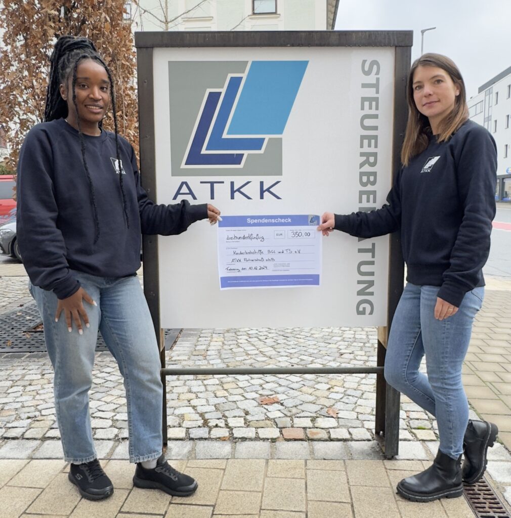 Zwei Mitarbeiterinnen der ATKK Partnerschaft mbB Steuerberatungsgesellschaft stehen mit einem Spendenscheck für die Kinderkrebshilfe BGL+TS vor ihrem Firmenschild.