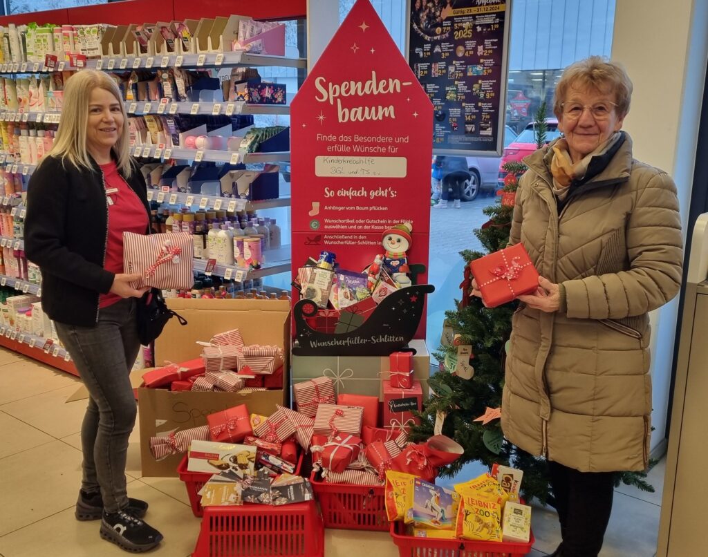 Spendenübergabe in der Laufener Rossmann-Filale: Christine Oberhofer, Leitung der Filiale Laufen und Rosmarie Baumgartner, 1. Vorsitzende Kinderkrebshilfe BGL+TS, stehen mit eingepackten Sachspenden vor dem Spendenbaum.