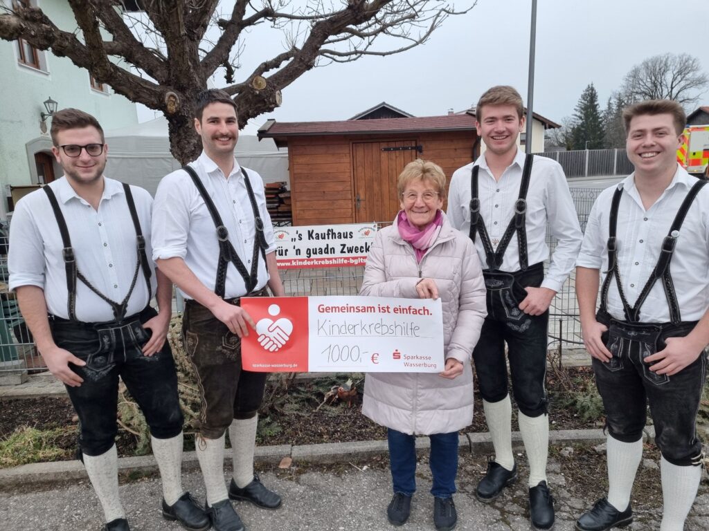 Vier Burschen des Burschenvereins Schnaitsee (alle in Lederhose gekleidet) überreichen vor dem Kaufhaus für'n guadn Zweck in Oberteisendorf ihren Spendenscheck an Rosmarie Baumgartner für die Kinderkrebshilfe BGL+TS.