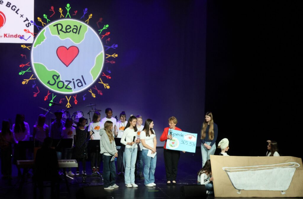 Auf einer Bühne, im Rampenlicht, nimmt Rosmarie Baumgartner von Schülerinnen der Walter-Mohr-Realschule Traunreut einen Spendenscheck entgegen. Oben sieht man das Logo von "Real Sozial": eine stilisierte Weltkugel mit Herz in der Mitte - rund um den Erdkreis herum reihen sich viele bunte Strichmännchen.