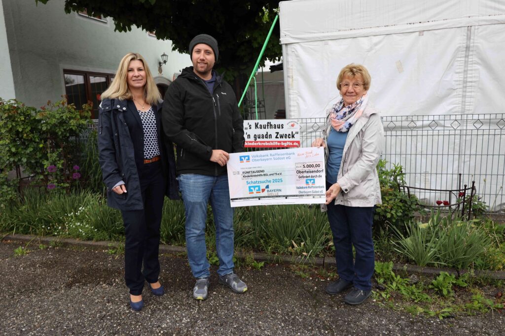Eine Vertreterin der Bayernwelle, der Gewinner Andi Dichtler und Rosmarie Baumgartner (1. Vorsitzende Kinderkrebshilfe BGL+TS) halten den Spendenscheck über 5.000 € - sie stehen auf dem Parkplatz vor dem Vereinsheim der Kinderkrebshilfe.